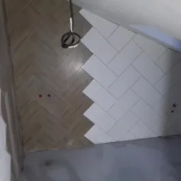 Fragment ściany łazienki w trakcie układania płytek w jodełkę, połączenie płytek drewnopodobnych i białych, widoczne wystające rury instalacji hydraulicznej.