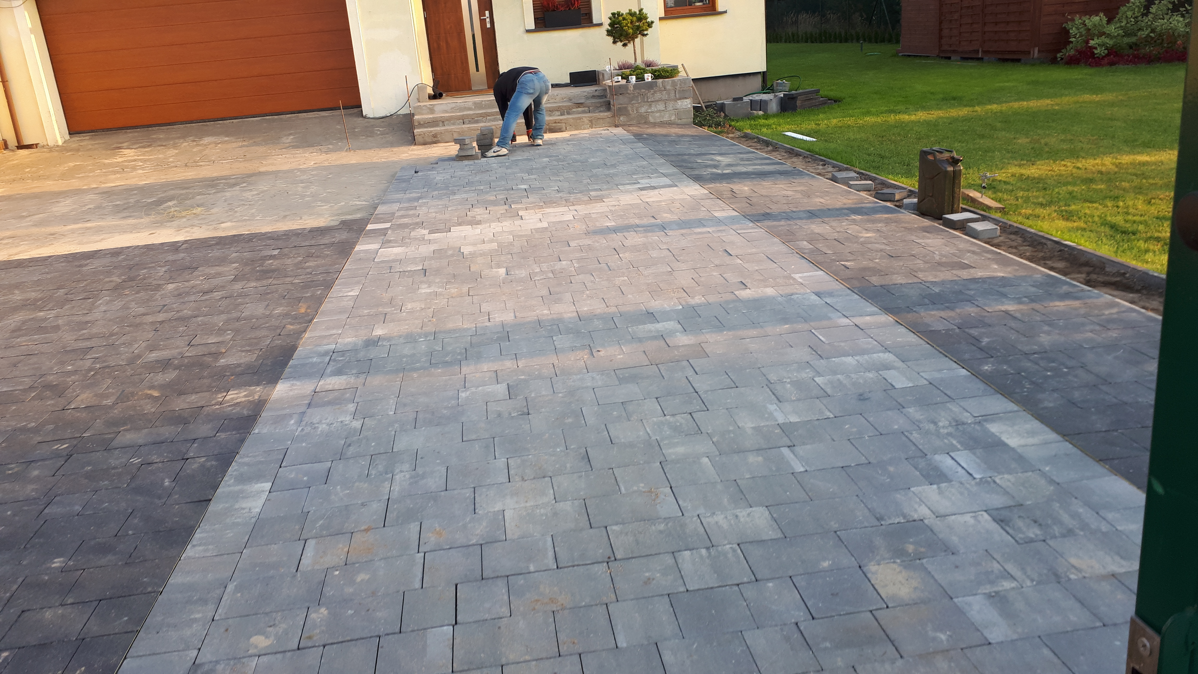 Układanie szarej kostki brukowej na podjeździe do domu jednorodzinnego, widoczny pracownik w trakcie montażu, część kostki już ułożona, reszta przygotowana do ułożenia.