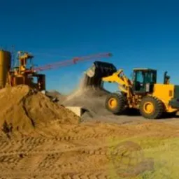 Żółta ładowarka czołowa wysypuje materiał z łyżki na pryzmę, w tle żółty silos i żuraw, błękitne niebo.