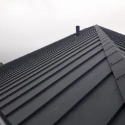 Nowy, ciemnoszary dach z blachy trapezowej, widoczny z dołu pod kątem, z kominkiem wentylacyjnym i fragmentem rynny.