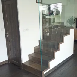 Nowoczesne schody z ciemnego drewna z szklaną balustradą, widok z dołu, drzwi w tle po lewej stronie.