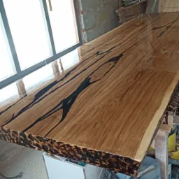 Surowy blat drewniany z żywicą epoksydową w warsztacie, widoczne niedoskonałości krawędzi i refleksy światła na powierzchni.