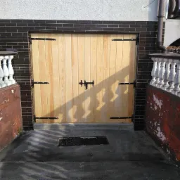 Jasne, pionowo ułożone deski tworzą bramę garażową z czarnymi zawiasami i prostym zamkiem, umieszczoną w ciemnej, ceglanej wnęce, z fragmentem balustrady po bokach.