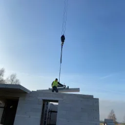 Montaż prefabrykowanego elementu stropu na budowie domu z bloczków silikatowych, z użyciem dźwigu i pracownika w kamizelce odblaskowej na tle błękitnego nieba.