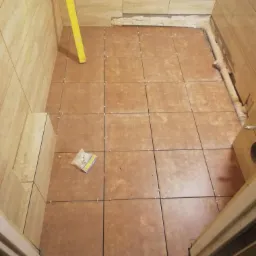 Brązowe płytki podłogowe w trakcie układania w małej łazience, widoczny poziomica i opakowanie krzyżyków dystansowych.