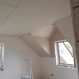 Montaż płyt na poddaszu: widoczne płyty na skośnym suficie, metalowa drabina, okno dachowe, wkrętarka i resztki materiałów na podłodze.