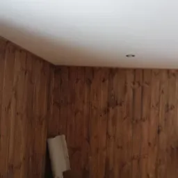 Pokój z boazerią w odcieniach brązu i białym sufitem z wbudowanym oświetleniem. Przy ścianie oparta rolka papieru lub folii.