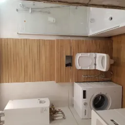 Nowoczesna łazienka z drewnianą okładziną, wiszącą toaletą z otwartą klapą, pralką i bojlerem, kabina prysznicowa w tle.