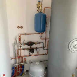 Instalacja centralnego ogrzewania z dużym szarym zbiornikiem, niebieskim naczyniem przeponowym i miedzianymi rurami, widoczne zawory, manometry i pompa obiegowa, ściany w kolorze złamanej bieli...