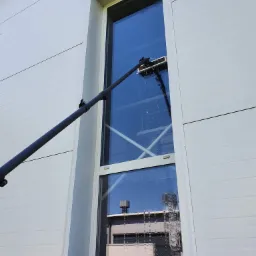 Mycie okna na wysokości przy użyciu teleskopowej lancy zakończonej szczotką i spryskiwaczem, widoczne krople wody na szybie, budynek z blachy trapezowej w tle.