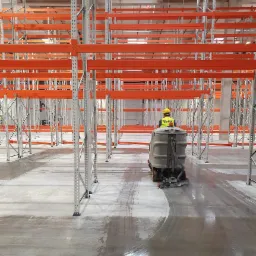 Operator w żółtym kasku i kamizelce, obsługuje maszynę czyszczącą posadzkę w magazynie z regałami paletowymi, widoczne mokre ślady po umytej powierzchni.