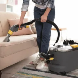 Kobieta w dżinsach czyści beżową kanapę za pomocą odkurzacza piorącego na tle jasnego wnętrza.