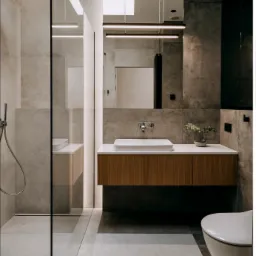 Nowoczesna łazienka z kabiną prysznicową typu walk-in, umywalką nablatową na drewnianej szafce i podwieszaną toaletą, wykończona płytkami w odcieniach szarości i beżu.