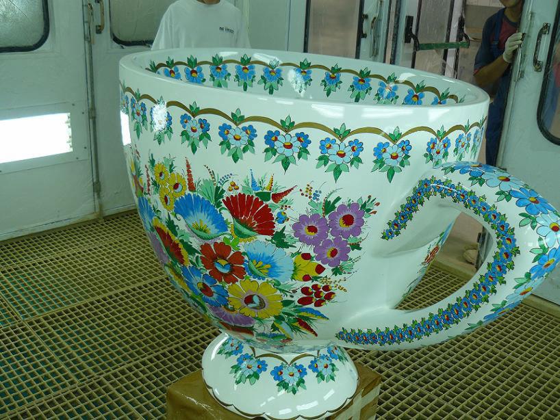 Ogromna, ręcznie malowana filiżanka w kwiaty, stojąca na kratownicy podłogowej. W tle osoby w białych i niebieskich ubraniach, drzwi.