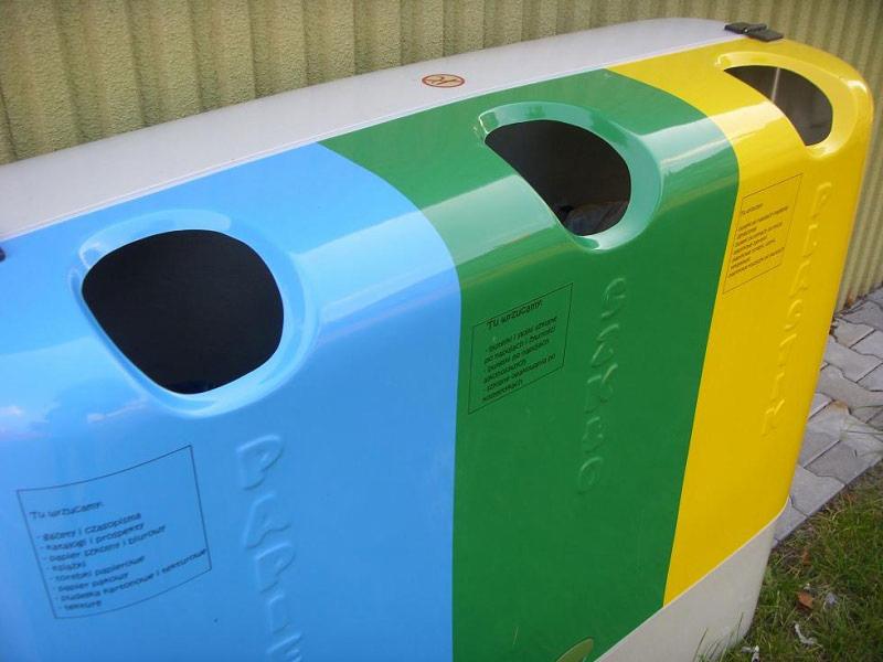 Kolorowy pojemnik do segregacji odpadów z trzema otworami: niebieski na papier, zielony na szkło, żółty na plastik. Widoczne napisy informacyjne.