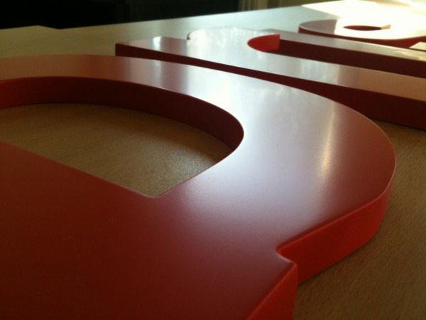 Czerwone, przestrzenne litery z tworzywa sztucznego, ułożone na jasnym blacie. Widoczne refleksy światła na gładkiej powierzchni liter.