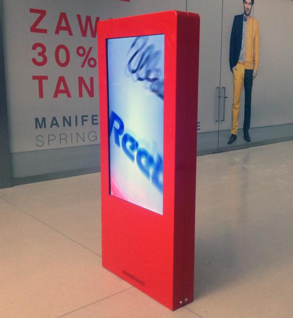 Czerwony, wolnostojący infokiosk z wyświetlaną reklamą Reebok w centrum handlowym, obok witryny sklepowej z manekinem.
