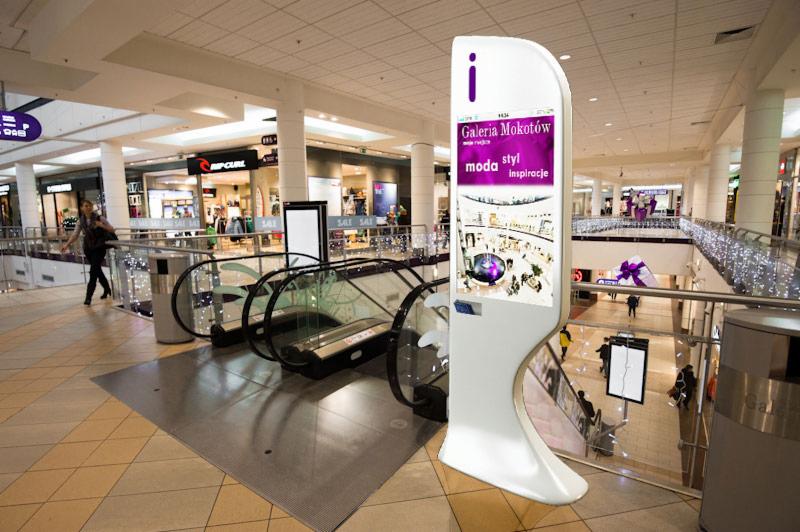 Interaktywny kiosk informacyjny 'Galeria Mokotów' z ekranem LCD, umieszczony w przestrzeni centrum handlowego, widoczny obok ruchomych schodów.