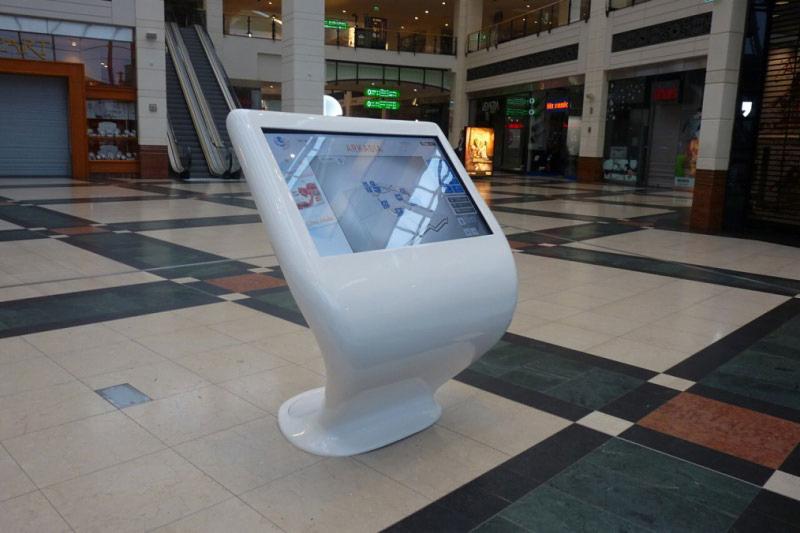 Interaktywny kiosk informacyjny z mapą centrum handlowego na ekranie, umieszczony na tle pasażu z eskalatorami i sklepami.