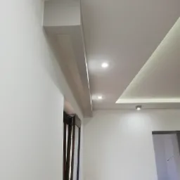 Nowoczesny sufit podwieszany z oświetleniem punktowym i listwą przysufitową, widok z dołu, białe ściany i częściowo widoczne okno przesuwne.