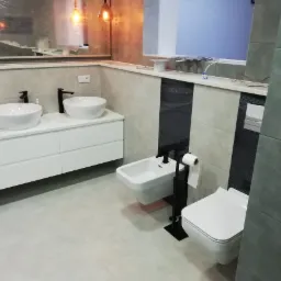 Nowoczesna łazienka z dwoma umywalkami nablatowymi na białej szafce, bidetem i toaletą, wykończona szarymi i białymi płytkami z czarnymi akcentami.