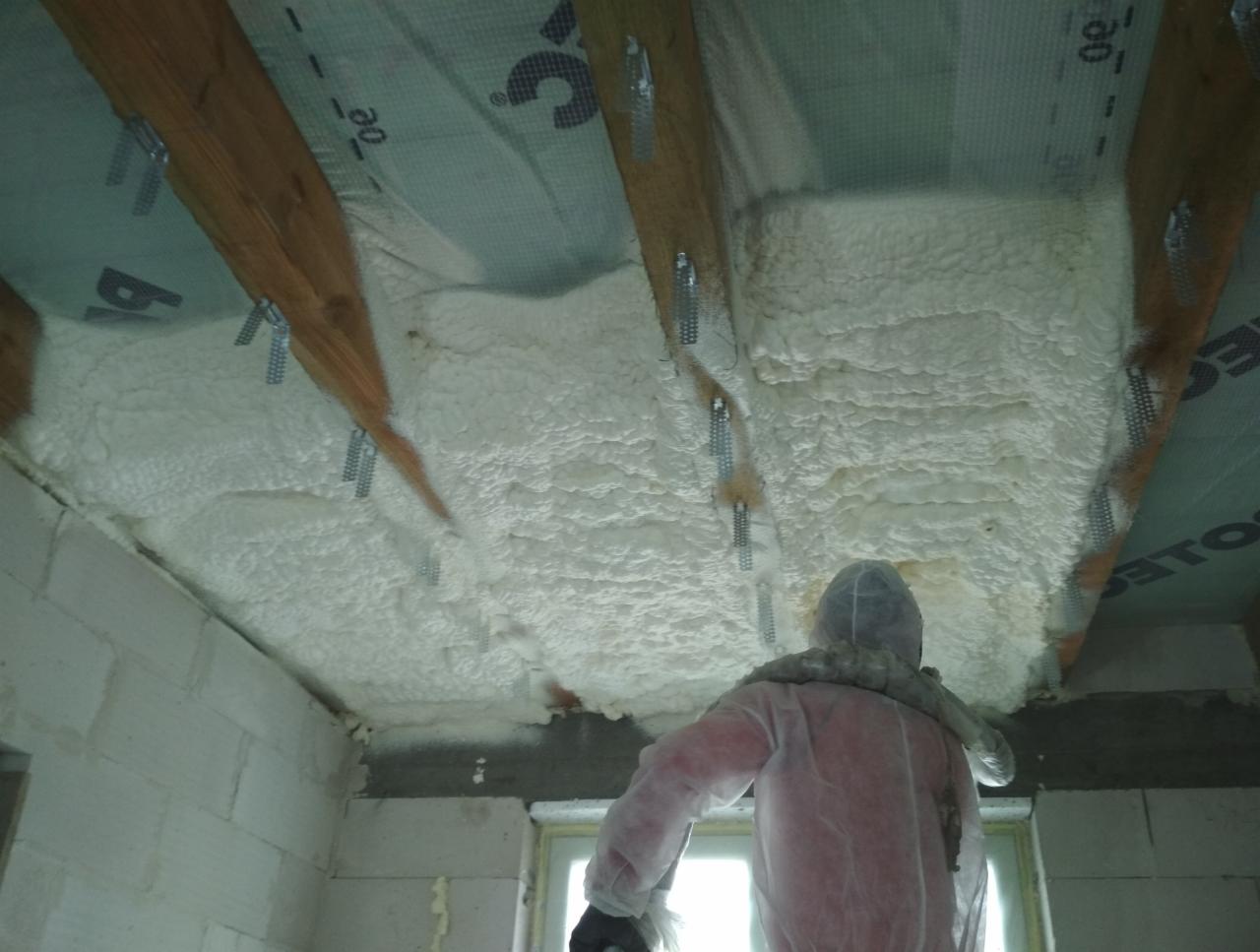 Pracownik w kombinezonie ochronnym aplikuje białą izolację na sufit poddasza z widocznymi drewnianymi belkami i metalowymi elementami mocującymi.