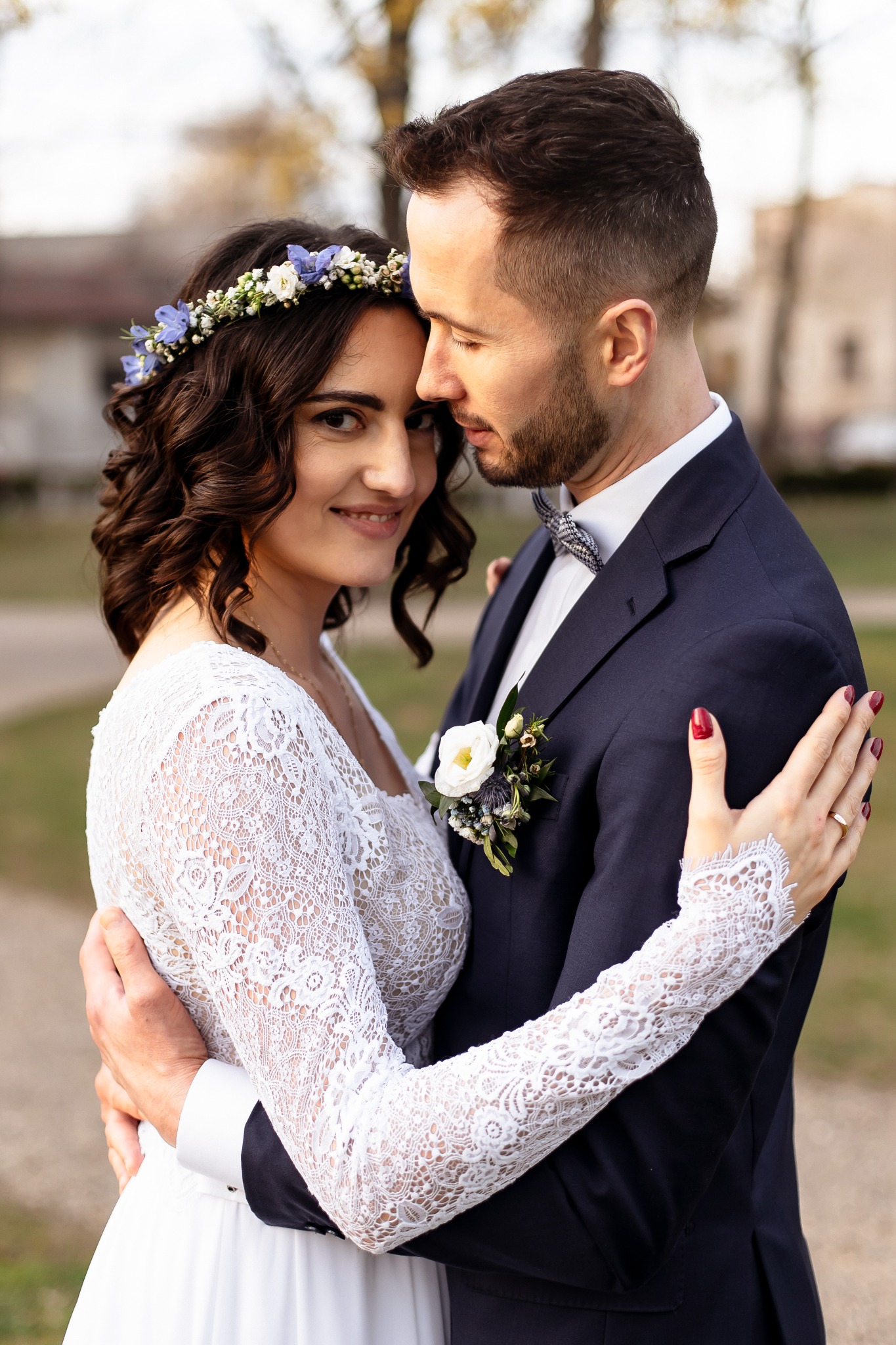 Uśmiechnięta panna młoda w koronkowej sukni i wianku z niebieskich kwiatów, obejmowana przez pana młodego w granatowym garniturze z muchą w kratę.