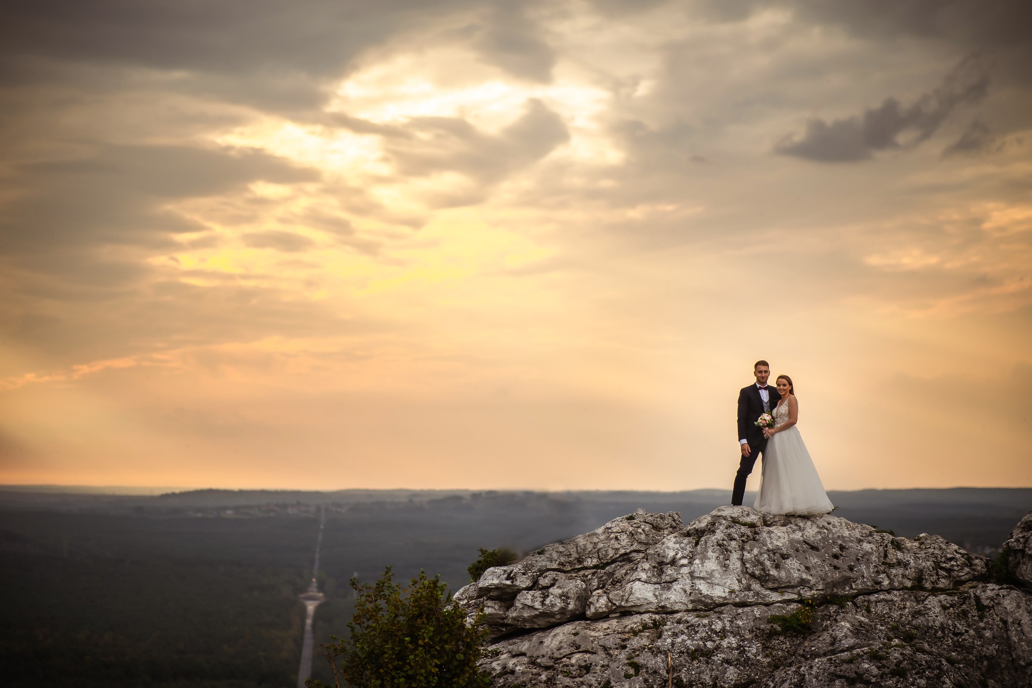 Para młoda w strojach ślubnych pozuje na skalistym wzniesieniu o zachodzie słońca, z widokiem na rozległy krajobraz i wieżę w oddali.
