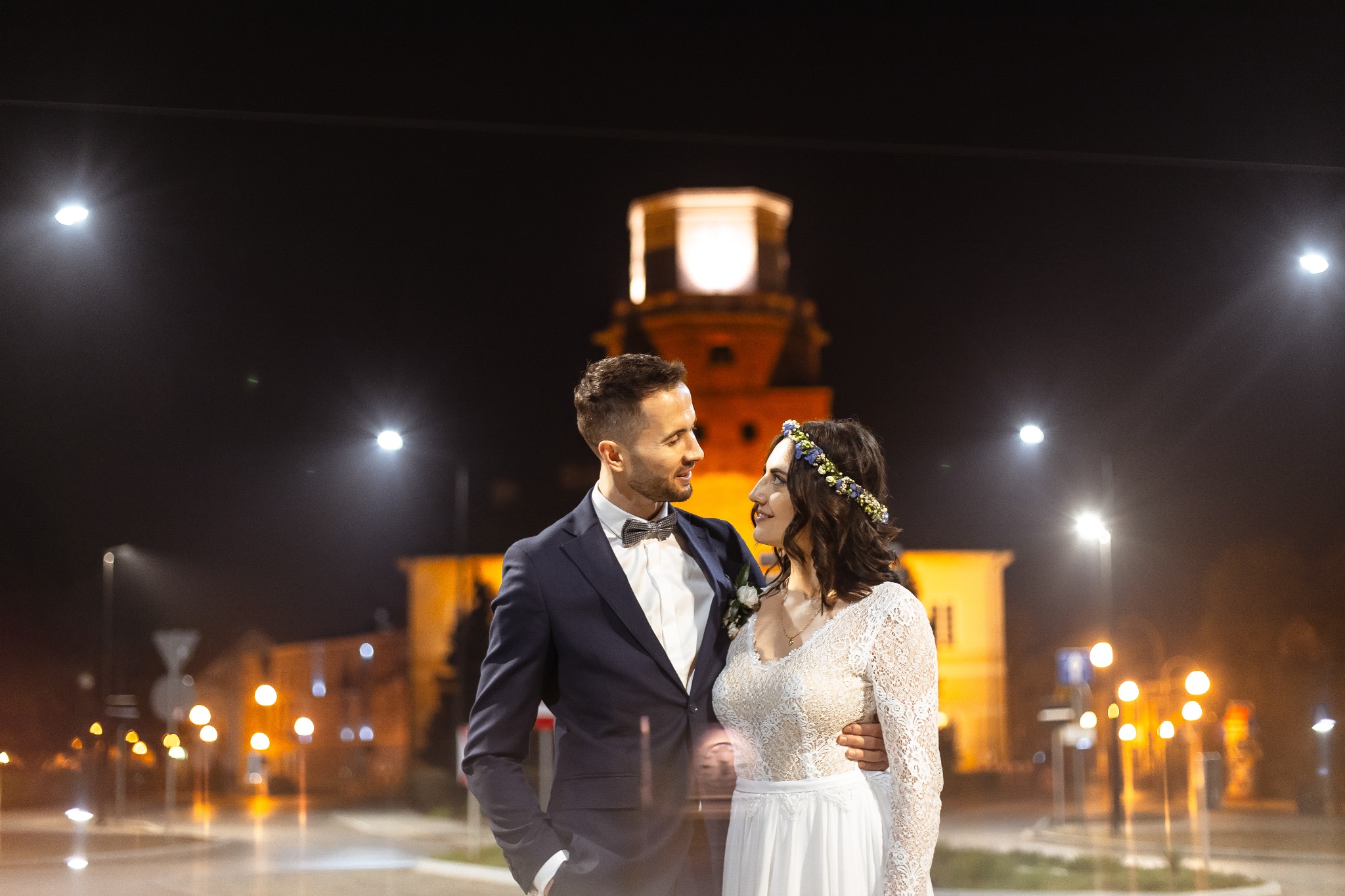 Para młoda w strojach ślubnych, ona z wiankiem na głowie, on w granatowym garniturze z muszką, obejmują się na tle nocnego krajobrazu z oświetlonym budynkiem w oddali.