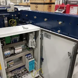 Otwarta szafa sterownicza z widocznymi elementami elektrycznymi: sterowniki PLC, bezpieczniki, przekaźniki, okablowanie. Na górze widoczny fragment konstrukcji stalowej w kolorze granatowym.