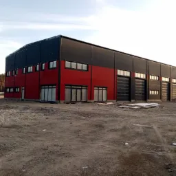 Nowoczesna hala przemysłowa z czarno-czerwonymi płytami warstwowymi, widok zewnętrzny, teren budowy w trakcie prac wykończeniowych, widoczne okna i bramy segmentowe.