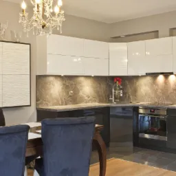 Nowoczesna kuchnia z białymi szafkami górnymi i ciemnymi dolnymi, marmurowym backsplashiem, oświetleniem punktowym pod szafkami i ozdobnym żyrandolem.