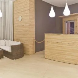 Recepcja z blatem imitującym drewno, z narożną sofą z rattanu i szklanym stolikiem kawowym, oświetlona wiszącymi lampami w kształcie łezki.