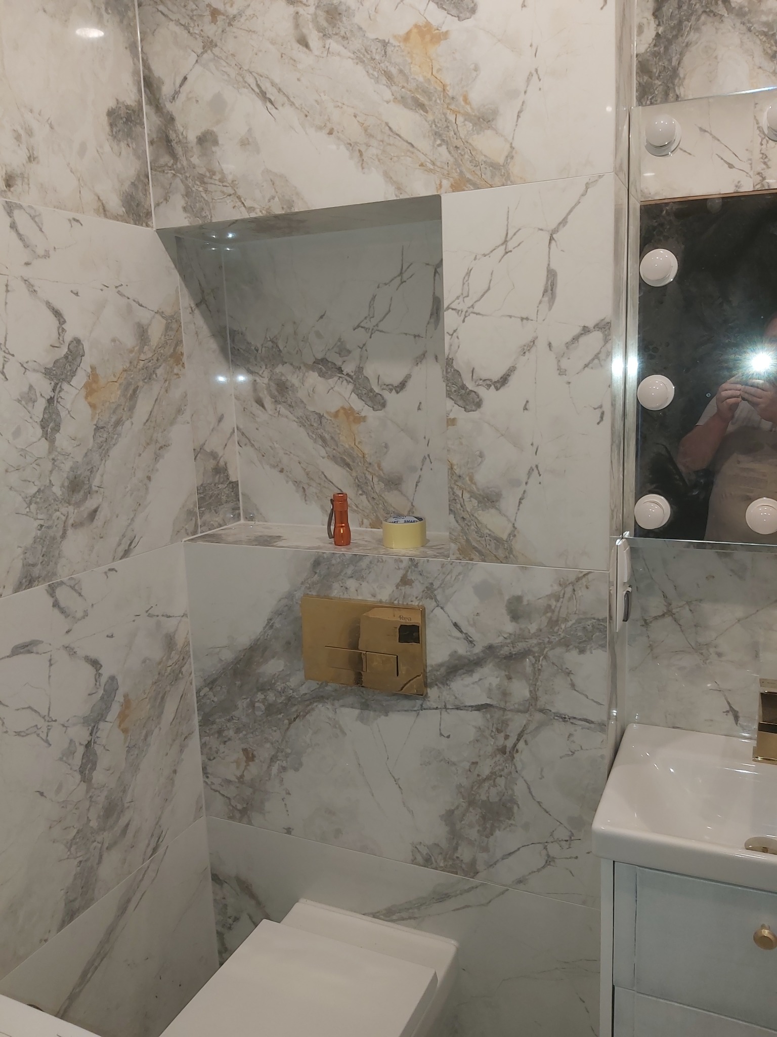 Wykończona łazienka z marmurowymi płytkami, podświetlanym lustrem i zabudowaną toaletą. Widoczny fragment instalacji i akcesoria, tworzące elegancki i nowoczesny wystrój.