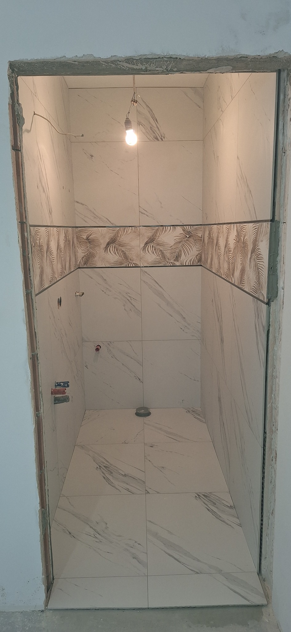 Wykończona wnęka prysznicowa z białymi płytkami imitującymi marmur i dekoracyjnym pasem z motywem liści palmowych. Widoczne przyłącza wodne i surowa ościeżnica.