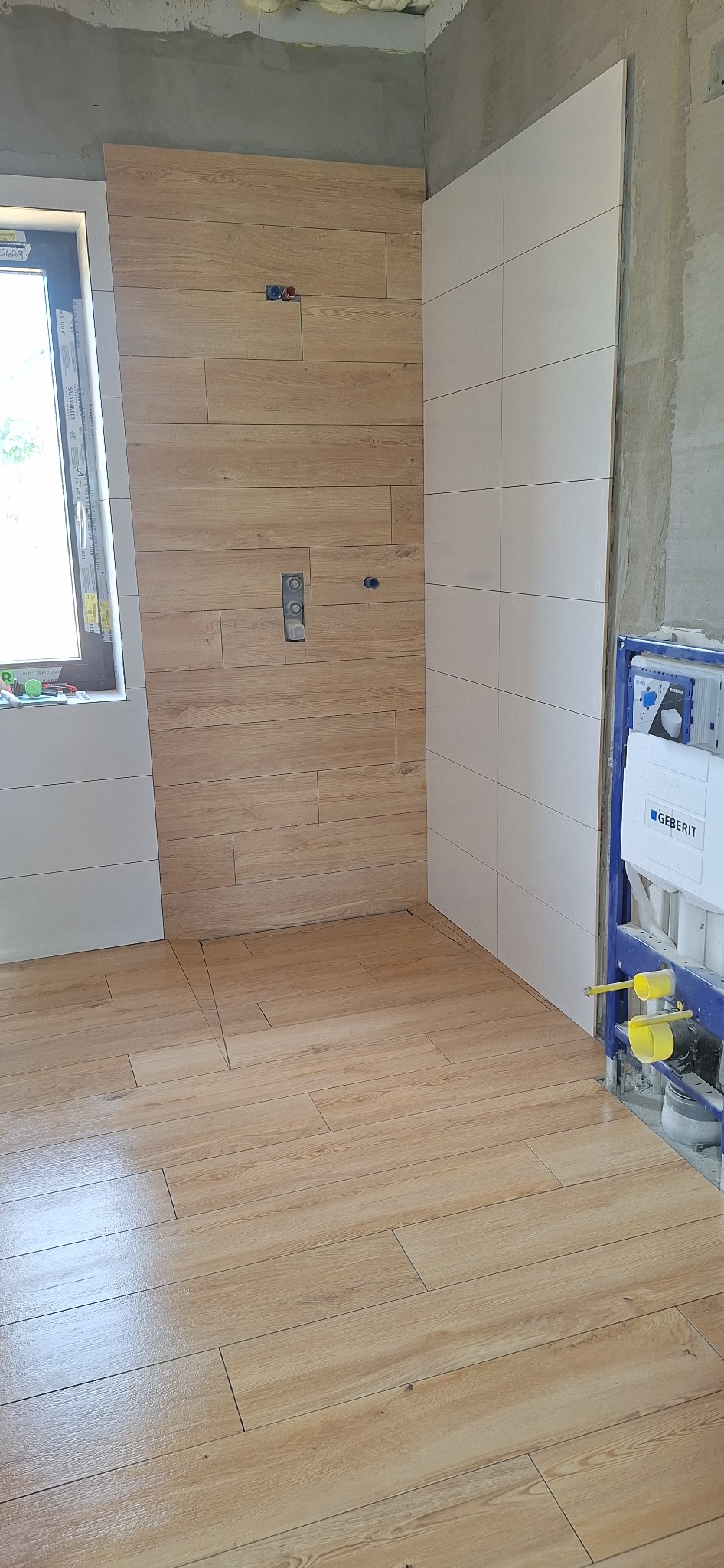 Układanie płytek imitujących drewno na podłodze i ścianie w łazience. Widoczna instalacja podtynkowa WC Geberit. Częściowo wykończone ściany.