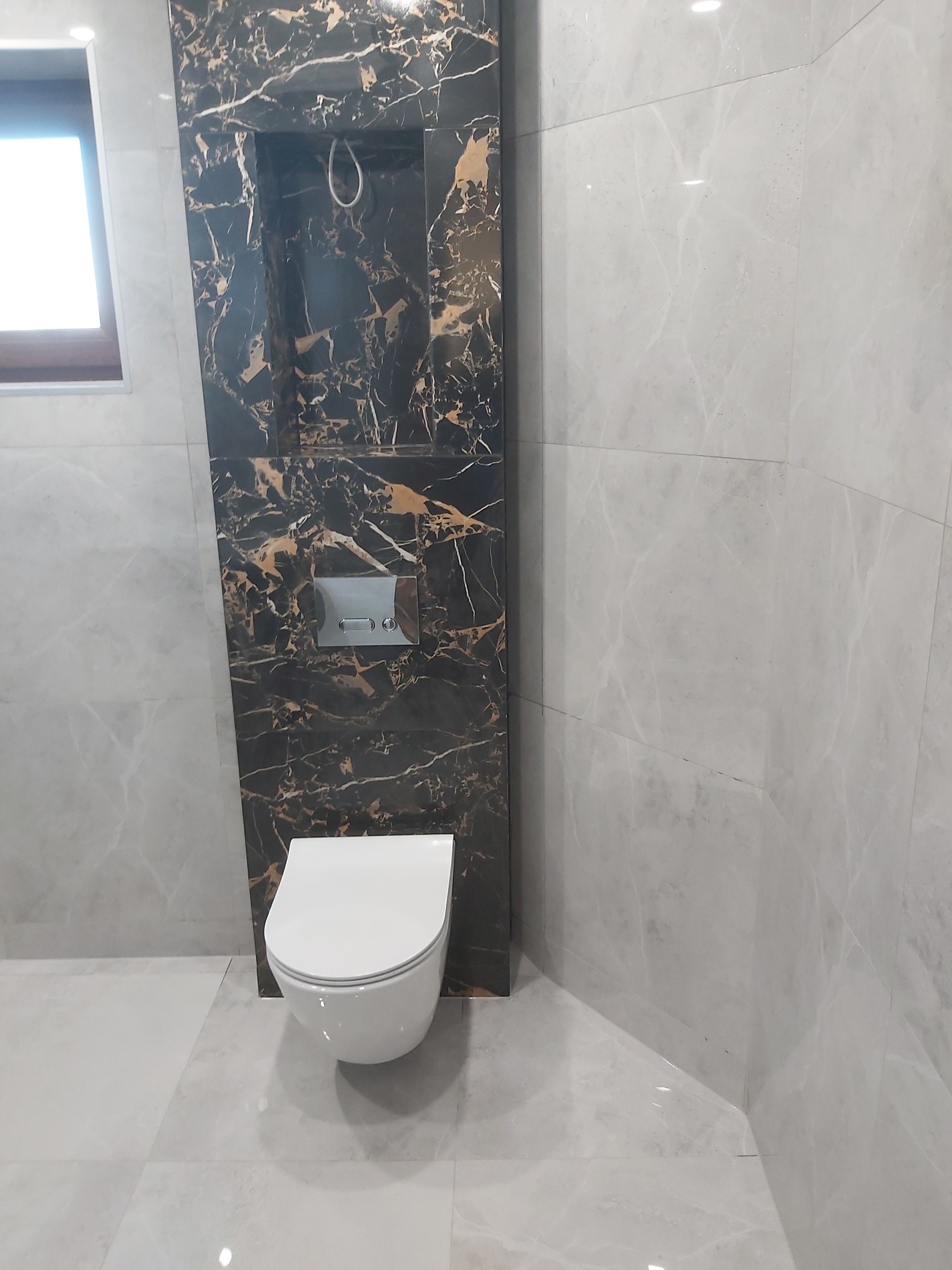 Nowoczesna łazienka z podwieszaną toaletą i czarną wnęką z marmurowym wzorem. Szare płytki na ścianach i podłodze. Minimalistyczny design, widoczne okno.