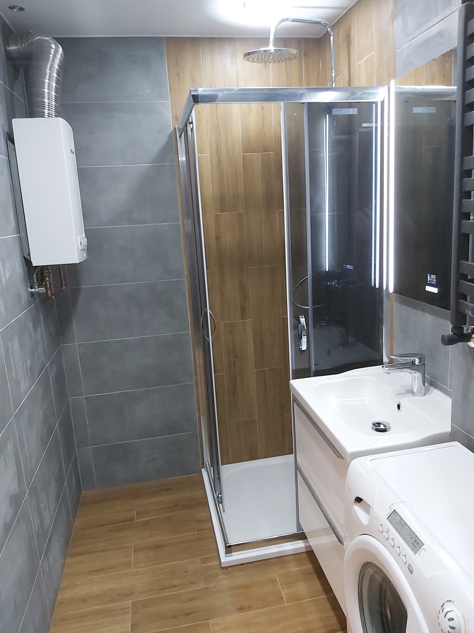 Nowoczesna łazienka z kabiną prysznicową typu walk-in, umywalką z szafką, pralką i grzejnikiem drabinkowym. Szare płytki ścienne i drewnopodobne płytki podłogowe.