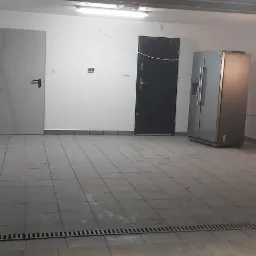 Przestronny garaż z szarą podłogą z płytek, białe ściany z dwiema parami drzwi, drabina, odkurzacz, półki na ścianie i osoba w kombinezonie malarskim.