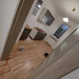 Wnętrze kuchni w trakcie remontu, widoczne panele podłogowe, częściowo zamontowane meble kuchenne, okno i lampa sufitowa, pomarańczowy kabel na podłodze, drzwi z szarym wykończeniem w pierwszym...