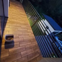 Sadowski Dawid - Ukończony montaż ogrodzenia z metalowych sztachet pionowych, widok z boku na drewnopodobną terakotę balkonu i wiaderko z resztkami zaprawy.