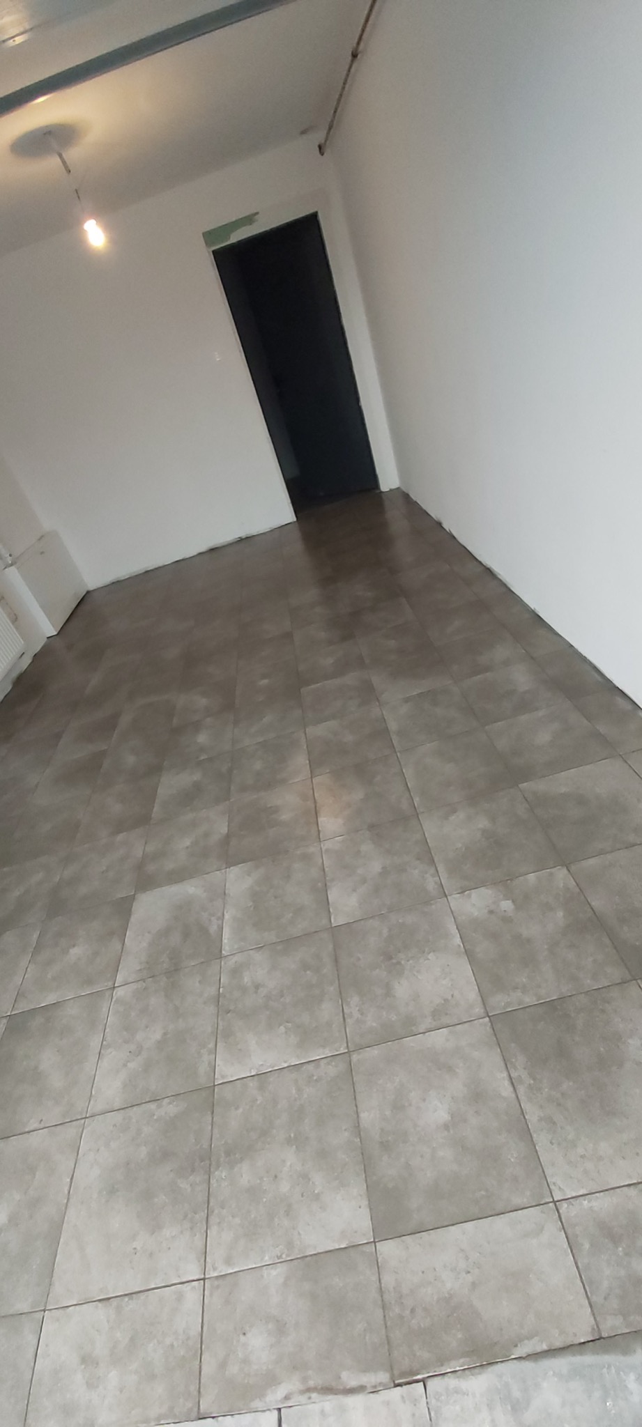 Wykończone wnętrze z ułożoną szarą terakotą podłogową, widoczna czarna ościeżnica drzwiowa i białe ściany, oświetlenie sufitowe w tle.