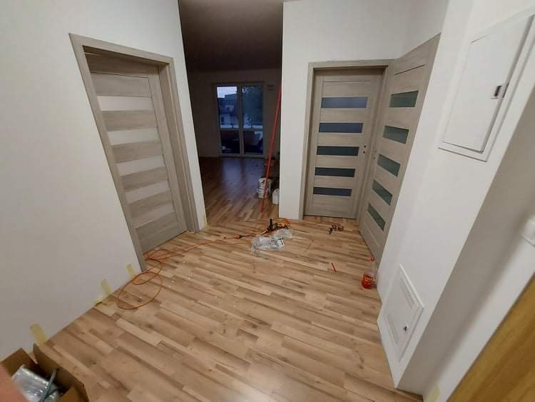 Wnętrze mieszkania w trakcie remontu, widoczne dwie zamontowane, nowoczesne, drewniane drzwi z poziomymi panelami, świeżo położona podłoga z paneli, przewody elektryczne na podłodze, kartony...