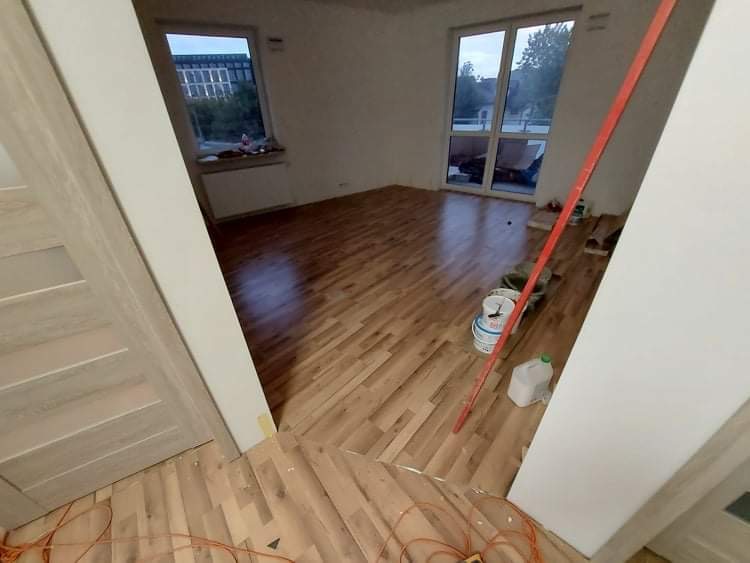 Wnętrze w trakcie wykańczania: świeżo ułożona, jasna podłoga panelowa w jodełkę, widoczne wiaderka z farbą, poziomica oparta o ścianę, okna z widokiem na budynek i zieleń.