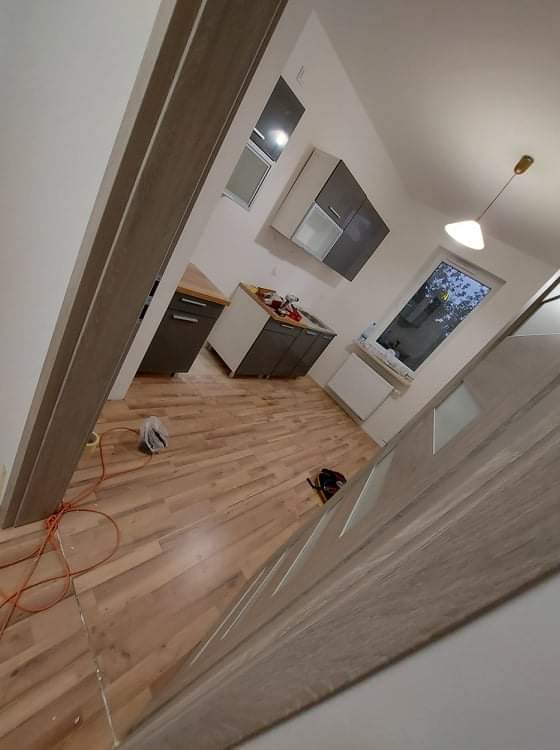 Wnętrze kuchni w trakcie remontu, widoczne panele podłogowe, częściowo zamontowane meble kuchenne, okno i lampa sufitowa, pomarańczowy kabel na podłodze, drzwi z szarym wykończeniem w pierwszym...