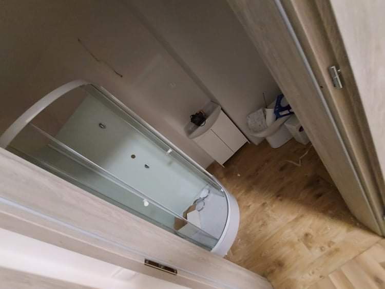 Nowoczesna kabina prysznicowa z zaokrąglonym szkłem w wykończonej łazience, widok przez otwarte, drewniane drzwi, jasne panele podłogowe, biała szafka umywalkowa w tle.