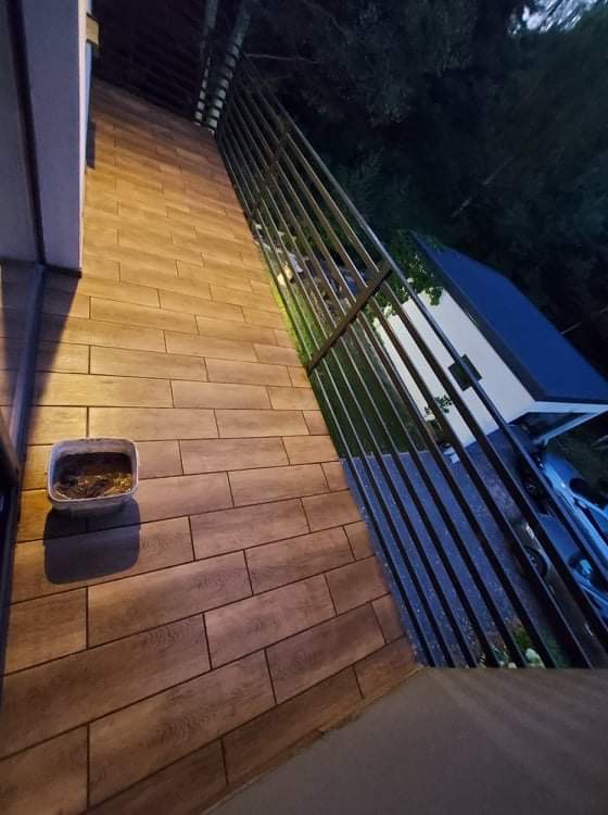 Ukończony montaż ogrodzenia z metalowych sztachet pionowych, widok z boku na drewnopodobną terakotę balkonu i wiaderko z resztkami zaprawy.