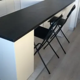 Wysoki blat kuchenny w kolorze czarnym, wsparty na białej podstawie, z dwoma czarnymi składanymi krzesłami, na tle jasnej podłogi i białych szafek.