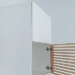 Biała szafka wisząca z otwartymi drzwiczkami, ukazującymi drewniane listwy wentylacyjne, zamontowana na jasnej ścianie.