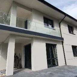 Nowoczesny dom z białymi ścianami, czarnymi ramami okiennymi i szklaną balustradą balkonu, otoczony zielenią i częściowo wyłożony szarą kostką brukową.